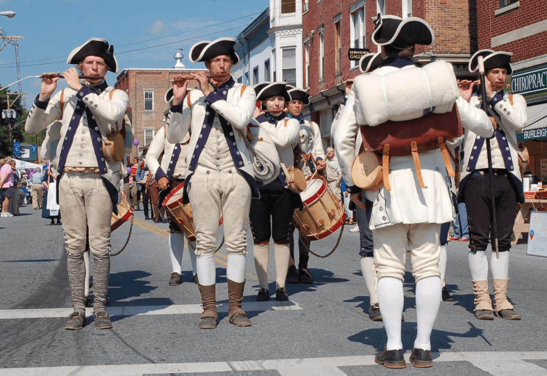 A group of people dressed in colonial-era costumes play flutes and drums while marching down the street during the Ticonderoga StreetFest parade, with brick buildings and spectators in the background.